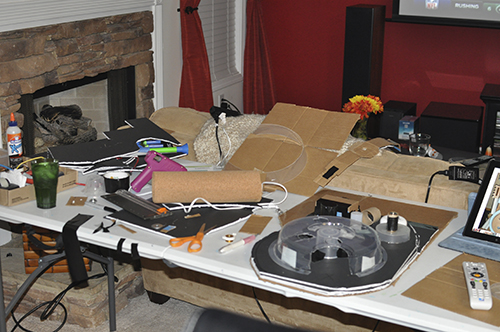 A messy work area of cardboard, glue gun, scissors, plastic and lots of other junky looking items. Creativity is messy!