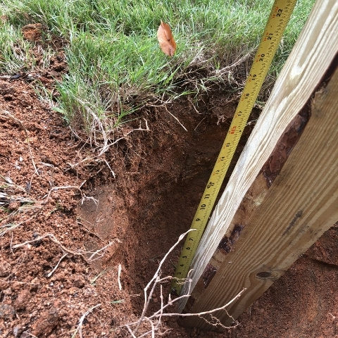 photo of a hole dug in the ground with tape measure showing 18 inches deep along with wooden post in the hole